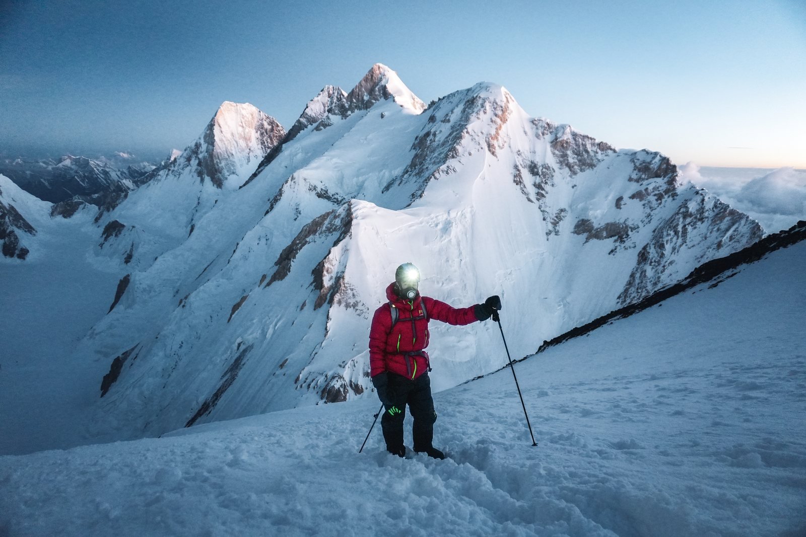 A climber in a red down jacket, headlamp on, on a snow slope at dawn, Karakoram summits behind